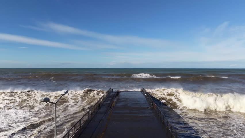 Slow motion footage of powerful ocean waves crashing forcefully against a concrete pier during rough sea conditions. Captured from a frontal perspective, the scene reveals dramatic splashes, thick sea foam, and turbulent water movement in exceptional detail. The slow motion effect enhances every impact, allowing the viewer to clearly see the strength, weight, and energy of the waves as they collide with the solid man-made structure.

This cinematic coastal scene highlights the raw power of nature and the constant interaction between the ocean and coastal infrastructure. The contrast between the moving water and the static pier creates a visually compelling composition that symbolizes resilience, endurance, and the unstoppable force of the sea. Natural daylight illuminates the textures of the waves, water droplets, and foam, adding depth, realism, and high visual quality to the footage.

Ideal for use in documentaries, cinematic intros, environmental storytelling, climate change visuals, weather-related projects, and nature-based video productions. This clip works well for themes such as stormy sea, coastal storms, marine energy, extreme weather, natural disasters, and the relationship between humans and nature. It can also be used as a dramatic background, opening shot, or visual transition in films, advertisements, travel videos, and social media content.

The immersive perspective places the viewer directly in front of the crashing waves, creating a strong emotional impact and sense of motion. This slow motion ocean footage is suitable for professional video projects requiring high-quality, dynamic, and atmospheric seaside visuals that convey power, movement, and intensity.