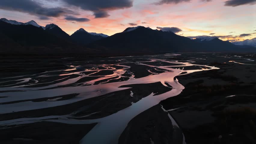 Aerial view of winding rivers reflecting a vibrant sunset, contrasted against the dark silhouettes of the mountains, Anchorage, Alaska, United States.