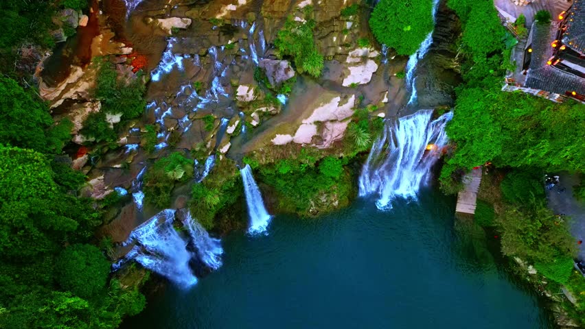 Aerial view of cascading waterfalls with lush greenery and traditional buildings, contrasting vibrant nature and architecture, Xiangxi Tujia, China.