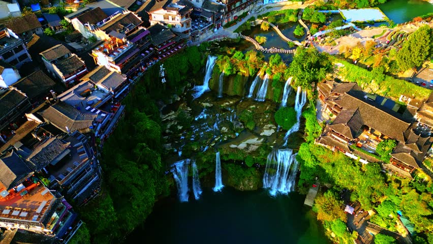 Aerial view of the Furong Ancient Town Waterfall cascading down the cliffside next to traditional buildings, Xiangxi Tujia and Miao Autonomous Prefecture, China.