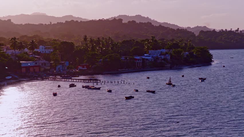 Sunrise over Miches in Dominican Republic, showing small fishing boats on calm coastal waters, with tropical hills, palm trees and quiet shoreline village glowing in warm morning light. Aerial orbit.