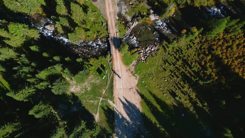 Overhead (top-down) aerial shot of a dirt road and a small bridge crossing a fast mountain stream. Two cyclists ride across the bridge, appearing tiny in a wide scenic landscape. Early autumn, sunny day.