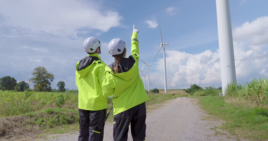 Two Caucasian engineer in safety jacket pointing toward wind turbine under blue sky discussing inspection teamwork concept of renewable energy project innovation professional maintenance slow motion