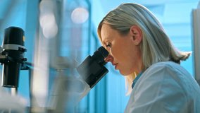 A focused female scientist in a white lab coat and blue gloves examines a sample under a microscope in a clean, well-lit modern laboratory. - Powered by Shutterstock - Get 15% off with code: PIKWIZARD15