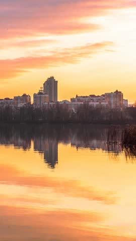 Sunset over Obolon embankment in Kyiv with modern residential skyline reflected in calm river water, dramatic clouds and warm golden light creating a peaceful urban evening atmosphere. Time lapse