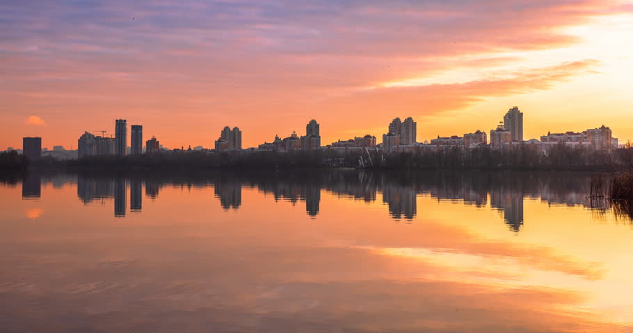 Sunset over Obolon embankment in Kyiv with modern residential skyline reflected in calm river water, dramatic clouds and warm golden light creating a peaceful urban evening atmosphere. Time lapse