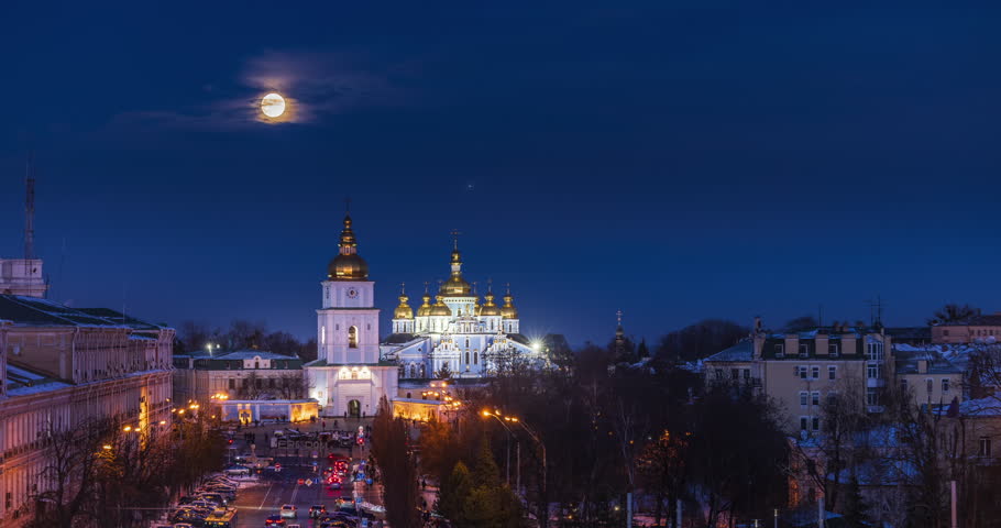 Winter evening at Sofia Square in Kyiv with the rising moon above St. Michael’s Golden-Domed Monastery, glowing domes, snowy streets, city lights, calm blue sky, historic urban atmosphere.