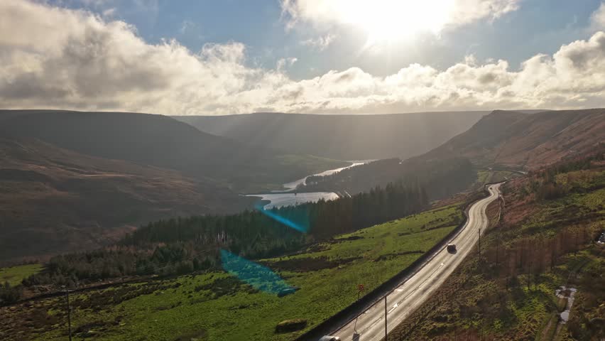 Aerial drone ascending frames Dovestones Reservoir with A635 road corridor visible green moorland conifer forest cumulus clouds fair weather excellent visibility Peak District Saddleworth, England