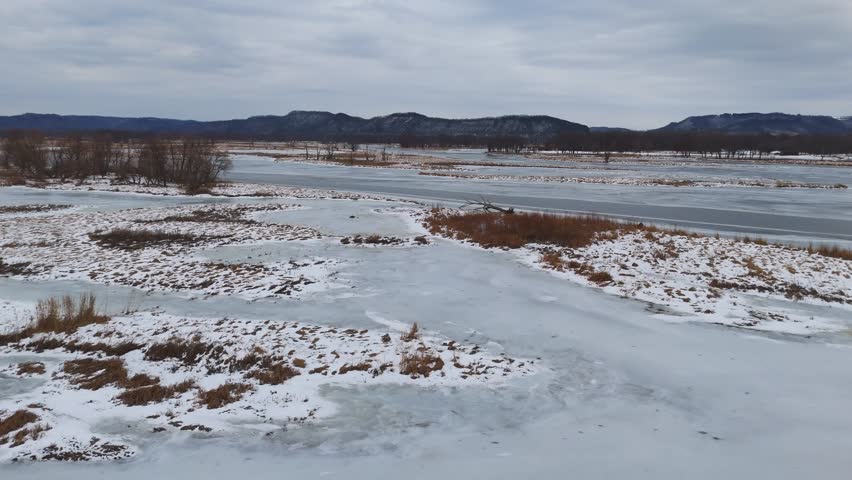 Winter stillness fills a partially iced wetland where dry grasses and bare trees stretch toward snowy hills on the horizon.