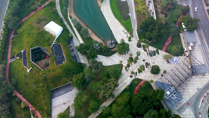 Top-down drone shot of Al Shaheed Park in Kuwait featuring a turquoise water feature, landscaped gardens, curved walkways, palm trees, and modern geometric structures in a detailed urban setting.