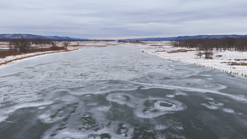 Shattered ice plates cover a wide river channel, framed by snow-dusted shores and leafless forests under pale winter light.