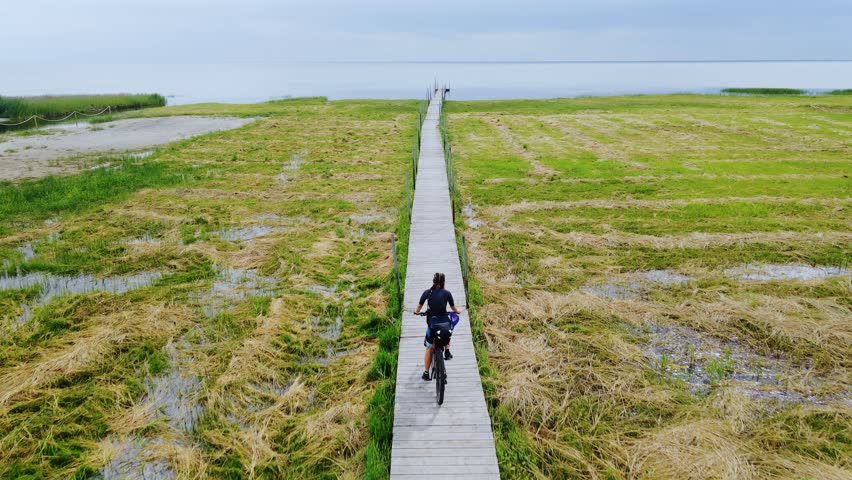 Female bikepacker cycles wooden boardwalk leading toward calm Baltic coastline