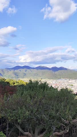 A stunning high-angle panoramic view of Kyoto city from a viewpoint within the Arashiyama forest. The footage shows the Katsura River winding through the urban landscape, bordered by lush greenery and autumn trees, with the distant mountains of the Kyoto basin under a blue sky with soft white clouds.