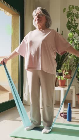 Active senior woman with grey hair exercising at home, performing a workout with elastic resistance bands on a yoga mat to improve her strength and mobility for a healthy lifestyle