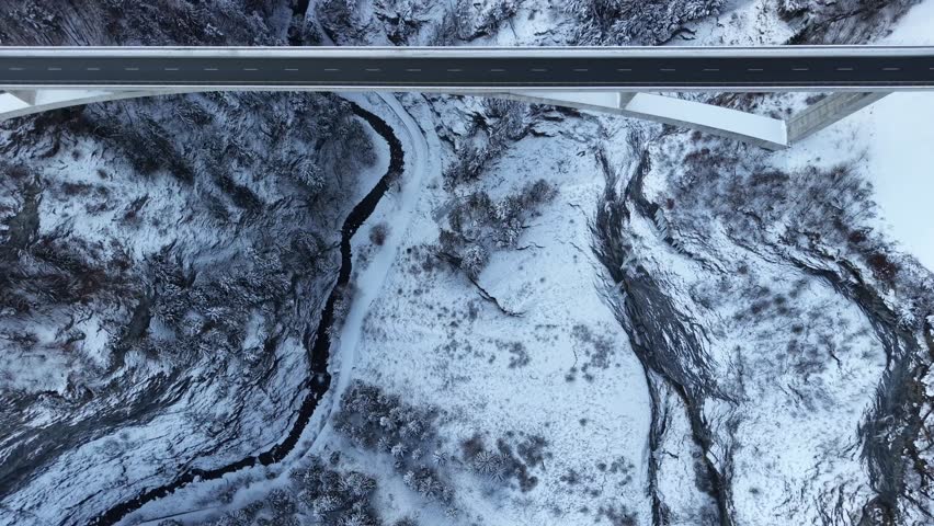 Top-down drone flight along a snowy gorge in Switzerland, showing the river below, passing under a bridge as a bus crosses, highlighting alpine winter scenery and infrastructure.