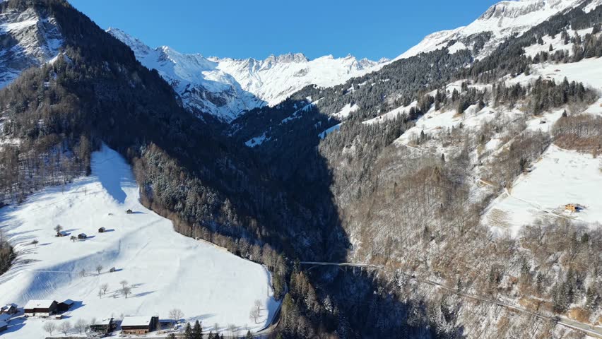 Aerial drone flight moving forward toward massive snow-capped mountains in Switzerland, with a forested midsection and a bridge crossing the valley below the peaks, highlighting alpine winter scenery and infrastructure integration.