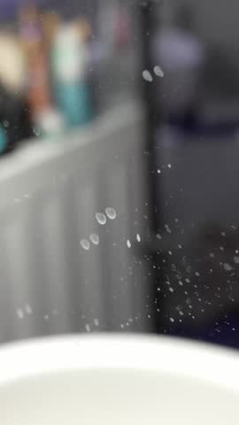 Macro shot of water spots and smudges on a bathroom mirror with a blurred sink and toiletries in the background, showing grime, residue, and poor cleaning.