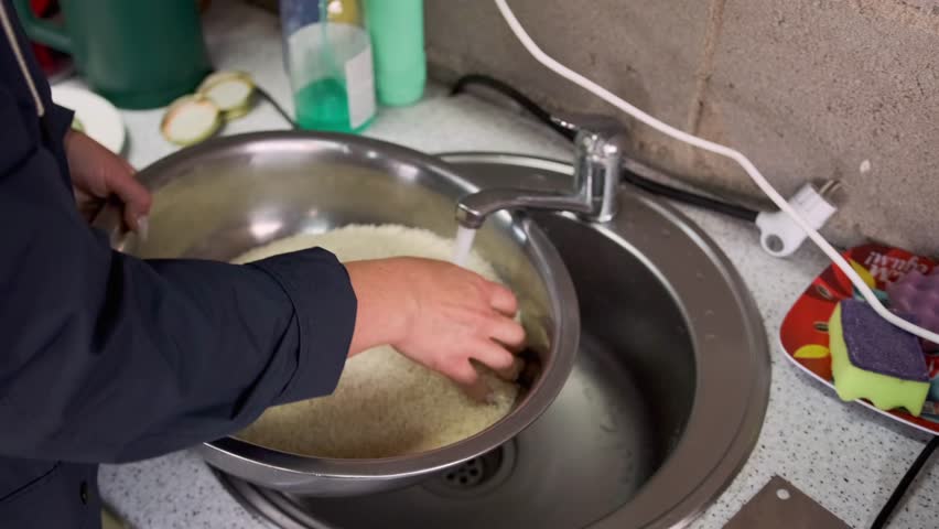 washing rice carefully, closeup of hands rinsing rice meticulously in sink, precise hands washing rice gently in metal bowl at sink, detailed closeup of hands carefully rinsing rice in metal bowl