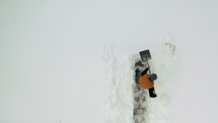 An aerial top view captures a man steadily making a clear path through thick, untouched snow after a heavy snowfall, showcasing his determined effort to reclaim the winter landscape.