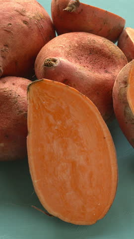 Whole and halved raw orange sweet potatoes on a blue background. Table spin. Vertical video.