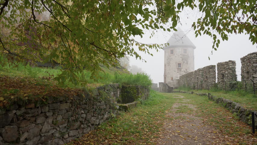 Ancient round stone tower and medieval fortification walls in thick fog during autumn. Path leads through historic castle grounds with misty atmosphere, mossy masonry and fall foliage.