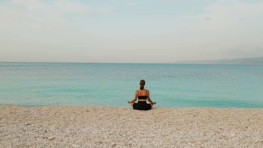 Woman meditating on pebble beach facing calm turquoise sea during peaceful morning