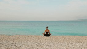 Woman meditating on pebble beach facing calm turquoise sea during peaceful morning - Powered by Shutterstock - Get 15% off with code: PIKWIZARD15