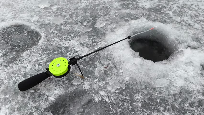 A close-up view of an ice fishing setup on a frozen lake. Bright green reel and fishing rod extend into a drilled hole in the ice. Crisp winter backdrop with ice formations and cold weather atmosphere