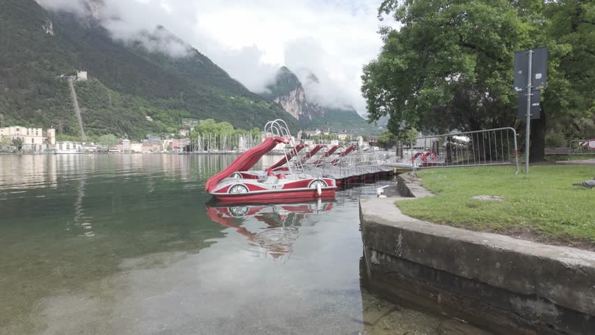 Riva del Garda, Italy - May 8, 2025 - Row of pedal boats with slides docked on a calm lake with mountains in the background