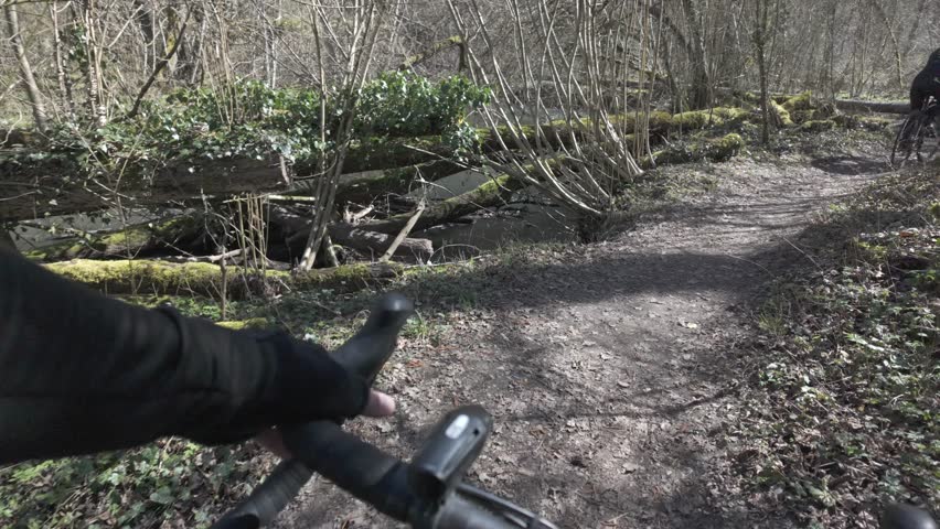 POV cycling through forest beside river following another cyclist on narrow dirt trail