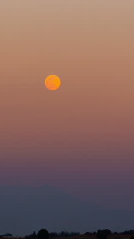 Timelapse of a stunning big orange full moon rising into the gradient twilight sky over a dark landscape