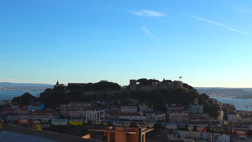 Sunlit panorama of Lisbon with São Jorge Castle on a hill and the Tagus River in the distance. Red rooftops and historic buildings fill the city below, filmed from a viewpoint platform.
