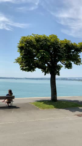 Peaceful summer day at Lake Zug, Switzerland. Two women sit on a bench at the waterfront promenade, enjoying the serene blue water and Alpine views under a clear sky. Authentic Swiss lifestyle.