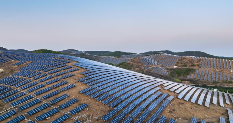 Aerial time-lapse of a mountain solar farm at dusk, showcasing rows of photovoltaic panels across rolling hills and a serene reservoir, highlighting renewable energy in harmony with nature.