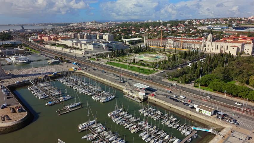 Aerial view of Lisbon Portugal showing marina with boats, Monument of the Discoveries, Tagus River waterfront, wide roads, parking areas and surrounding urban buildings next to Palace of Ajuda