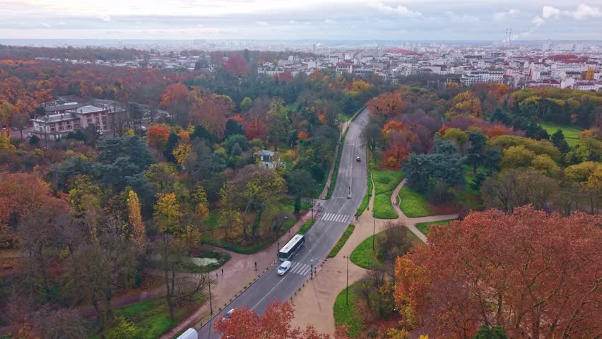 Aerial drone panorama reveals Avenue de Gravelle bisecting Bois de Vincennes autumn woodland with golden foliage, Château de Vincennes skyline and Paris suburbs beyond, France.