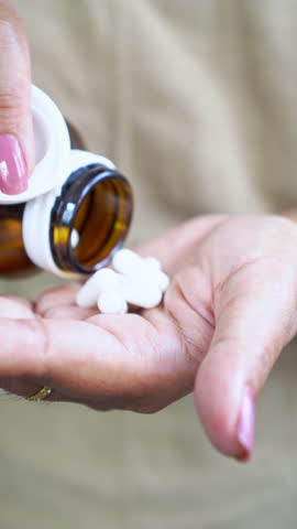 Vertical close up of a woman's hand holding a medicine bottle while taking vitamin pills and supplements like probiotics or magnesium to support her daily health, wellness, and medical wellbeing