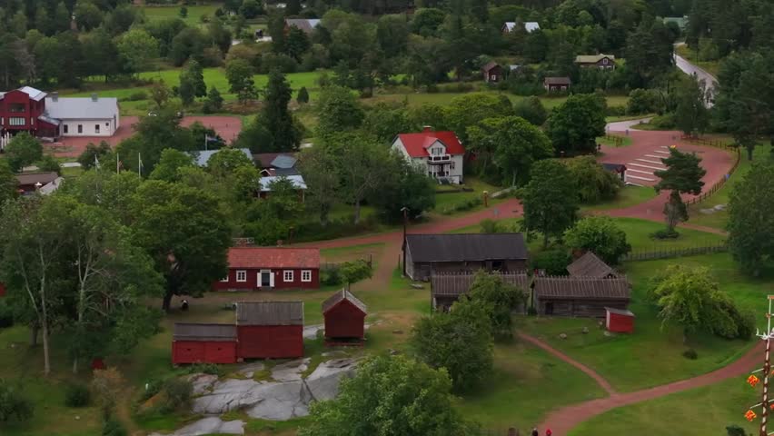 "Lush green trees surround a quaint Finnish village with traditional red-and-white buildings, and a winding gravel road meandering through the peaceful landscape."