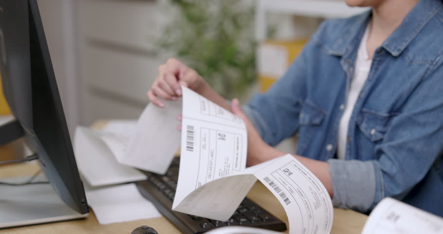Asian female entrepreneur pulls long shipping labels from printer while checking customer orders on computer during e-commerce startup daily work inside home warehouse storage area