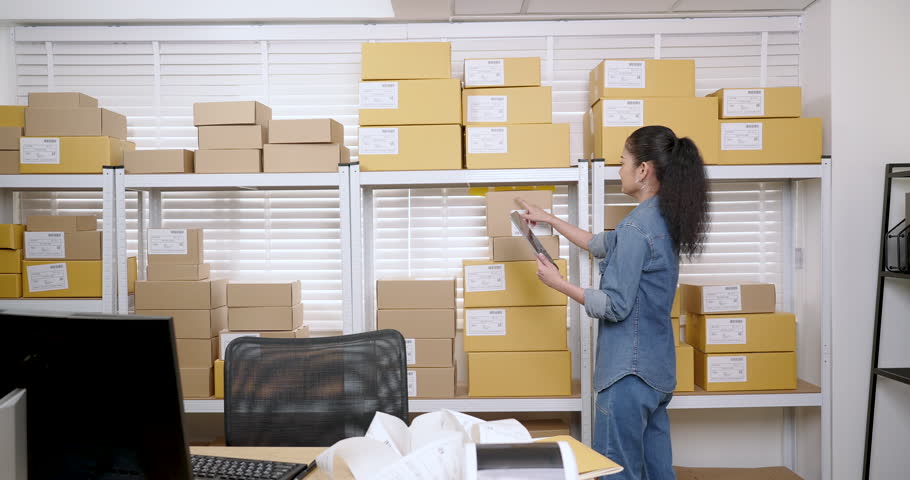 Asian woman entrepreneur walks across storage room, reviews inventory count, rearranges online shopping parcel box piles, ensures online order management accuracy for small business supply chain