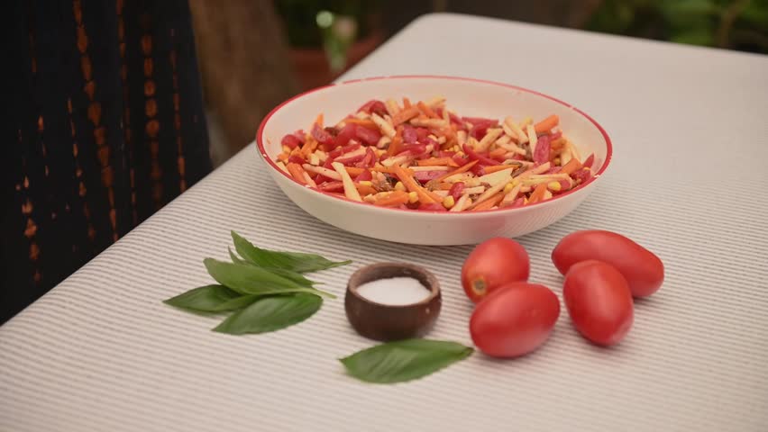Close-up video of a woman's hands mixing a fresh summer salad with chopped vegetables in a bowl. On the table there are tomatoes, fresh basil and salt, suggesting simple and healthy home cooking. Natu