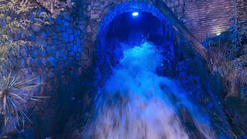 At night, a radiant man made waterfall cascades down from a stone archway, captivating viewers with its enchanting glow of bright blue lights shimmering on the water and the lush foliage around it
