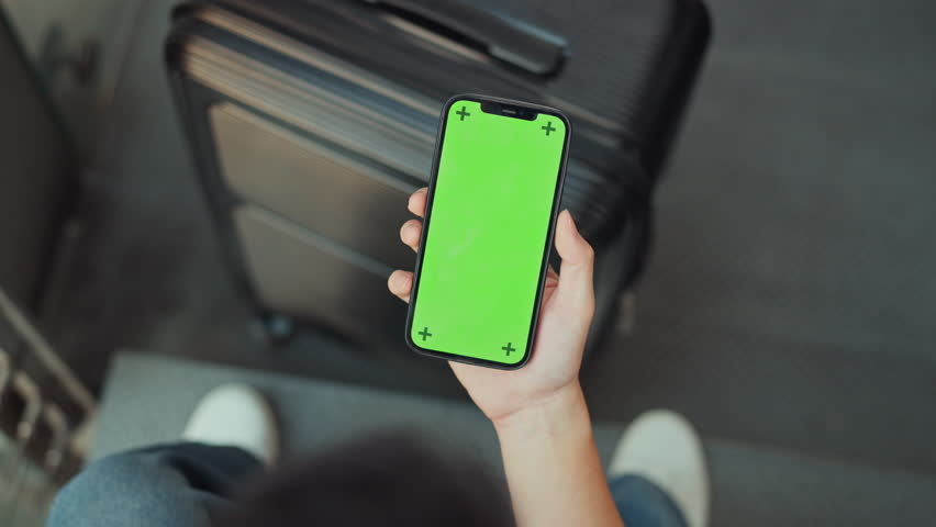 Top view POV of a traveler holding a smartphone with a green screen and tracking markers over a black suitcase while waiting in an airport or transit station