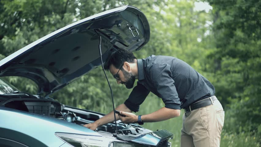 Driver Trying To Fix Engine Problems Or Checking Oil Level During Road Trip Emergency. Driver Inspecting Open Hood Of Broken Car. Troubleshooting Vehicle Failure. Automotive Accident Technical Support