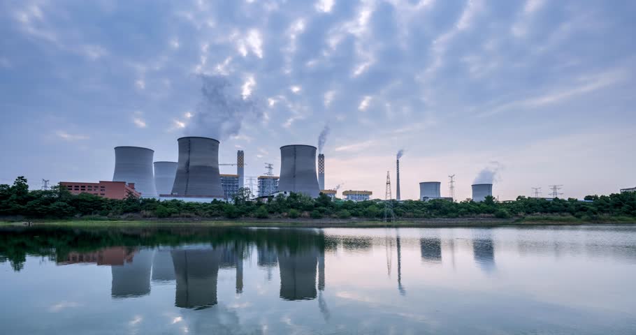 Time-lapse of a coal-fired power plant at dusk, with cooling towers emitting steam reflected in calm lake waters against a sunset sky.  