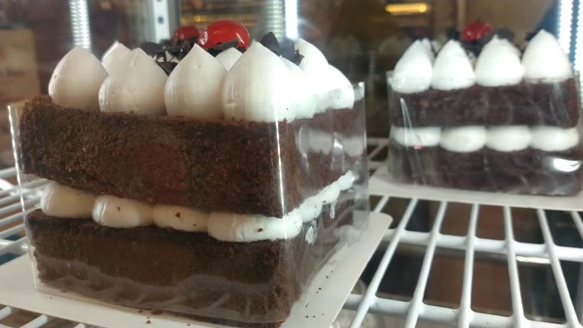 A layered chocolate cake topped with strawberries, whipped cream, and chocolate chips is displayed in a glass display case at a bakery.