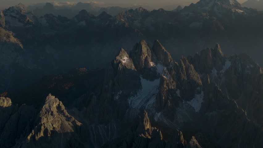 Cinematic aerial view of the Dolomites in Italy, rugged alpine peaks glowing at dawn, dramatic cliffs and deep shadowed valleys creating a serene high-altitude mood
