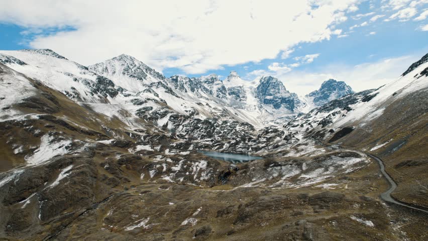 Breathtaking drone footage of the rugged, snow-dusted Andes mountains at Pico Austria. A remote glacial lake sits nestled among the peaks under a clear blue sky in the Bolivian Altiplano.