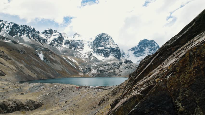 A sweeping aerial pan of the turquoise Chiarkhota glacial lake at the foot of jagged Andean peaks. The high-altitude majesty of the Pico Austria region in Bolivia.
