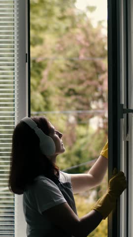 A woman wears headphones while cleaning a window with yellow gloves during daylight.
